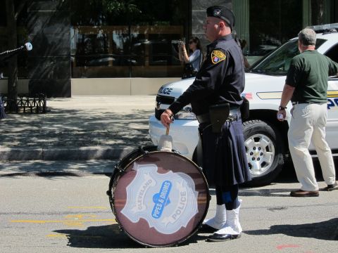 1991 Ford LTD Crown Victoria - uniformed drummer in a kilt rests a bass drum labeled Cleveland Police Pipes & Drums on the street beside a police SUV.