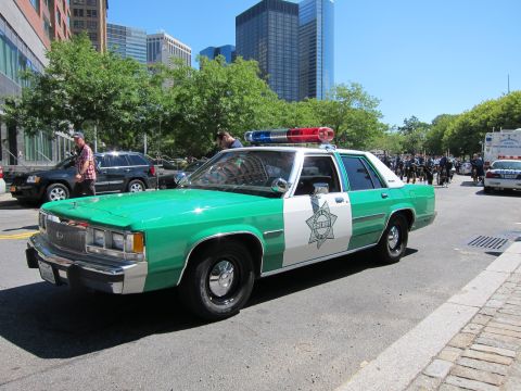 1991 Ford LTD Crown Victoria - green-and-white vintage Ford police sedan with a roof light bar and star decal sits on a downtown street near skyscrapers.