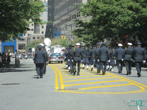 1991 Ford LTD Crown Victoria - uniformed honor guard and band march down a city street past TV satellite trucks and tall buildings.