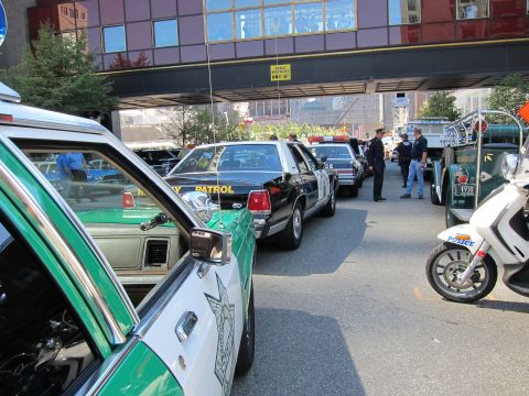 1991 Ford LTD Crown Victoria - lineup of vintage police cars, including green-and-white and black sedans, parks under a building overpass with a police motorcycle nearby.