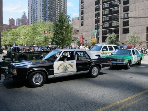 1991 Ford LTD Crown Victoria - black-and-white Highway Patrol sedan sits in a row with a white police car and a green-and-white sedan on a sunny city street.