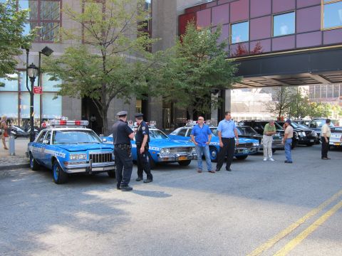 1991 Ford LTD Crown Victoria - Two blue vintage police sedans with roof lights are parked outside a building as officers and onlookers stand nearby.