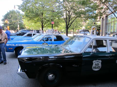 1991 Ford LTD Crown Victoria - black-and-green vintage City of New York Police sedan with a shield decal sits beside a blue police car on a shaded street.