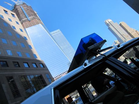1991 Ford LTD Crown Victoria - blue emergency light tops a vehicle, framed against a tall glass skyscraper still under construction and surrounding office towers.
