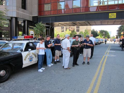 1991 Ford LTD Crown Victoria - group of people chats beside a black-and-white Highway Patrol sedan parked under a building overpass marked Max Height 14 ft 6 in.