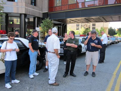 1991 Ford LTD Crown Victoria - Several people stand talking among a row of parked vintage police cars beneath a covered building overpass.