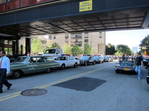 1991 Ford LTD Crown Victoria - row of vintage police cars, including a green Stratford Police sedan, lines a street under a building overpass with a crowd beyond.