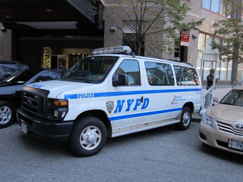 1991 Ford LTD Crown Victoria - white Ford NYPD passenger van with blue striping is parked beside a tan Toyota sedan along a building-lined city street.