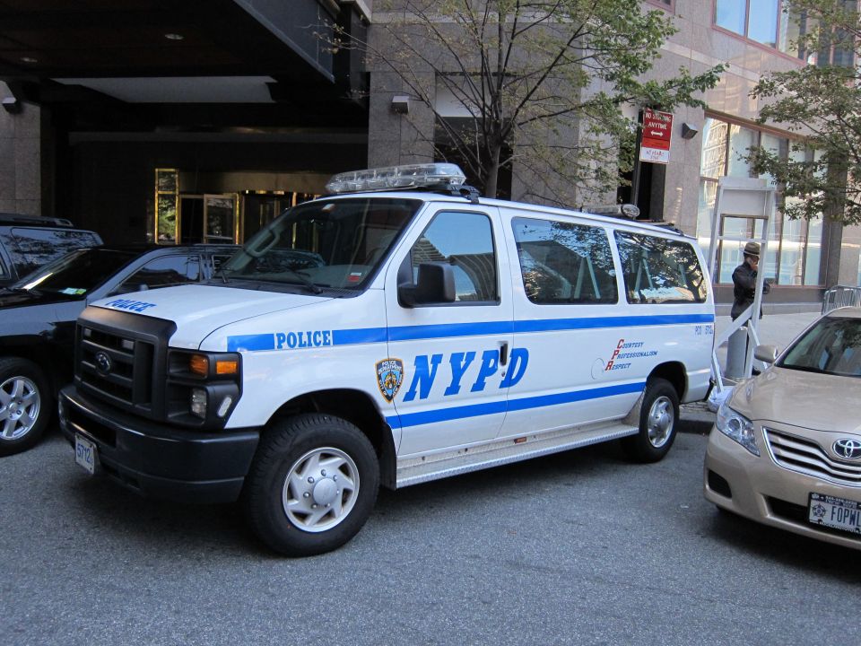 White Ford NYPD passenger van parked beside a tan Toyota sedan on a city street