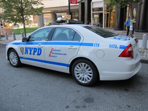 1991 Ford LTD Crown Victoria - white Ford Fusion NYPD patrol sedan with blue striping and roof light bar is parked along a city street.