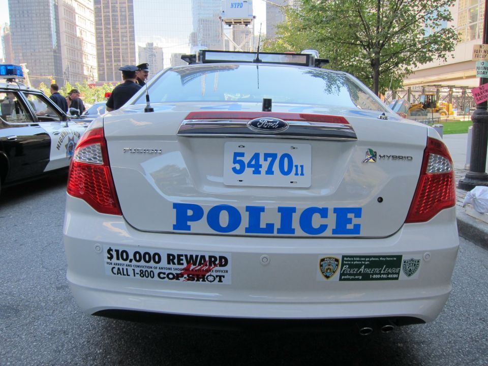 Rear of a white Ford Police hybrid sedan with plate 547011 and $10,000 reward decal