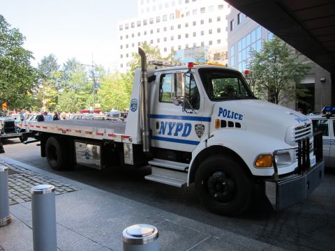 1991 Ford LTD Crown Victoria - white Sterling flatbed tow truck lettered NYPD POLICE sits on a stone plaza with office towers behind it.