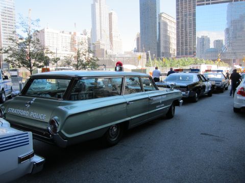 1991 Ford LTD Crown Victoria - 1960s green station wagon marked Stratford Police is parked in a row of police vehicles with high-rise buildings behind.
