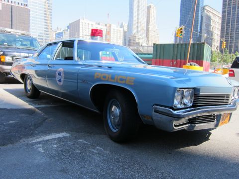 1991 Ford LTD Crown Victoria - light-blue 1970s four-door Police sedan with a roof light sits in a lot with high-rises and shipping containers behind.