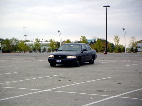 1999 Ford Crown Victoria - A dark sedan is parked in a large, empty parking lot under an overcast sky.