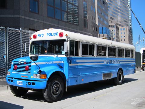 1991 Ford LTD Crown Victoria - blue NYC Police bus built on an International chassis is parked beside an office building downtown.