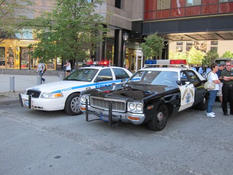 1991 Ford LTD Crown Victoria - white Ford Crown Victoria police car sits next to a 1970s black-and-white Highway Patrol cruiser outside a building.