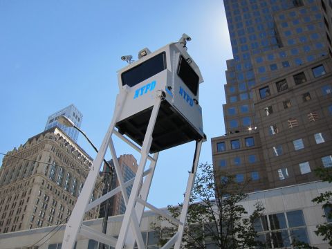 1991 Ford LTD Crown Victoria - white elevated NYPD surveillance booth mounted on a tall steel frame rises against office towers and clear sky.
