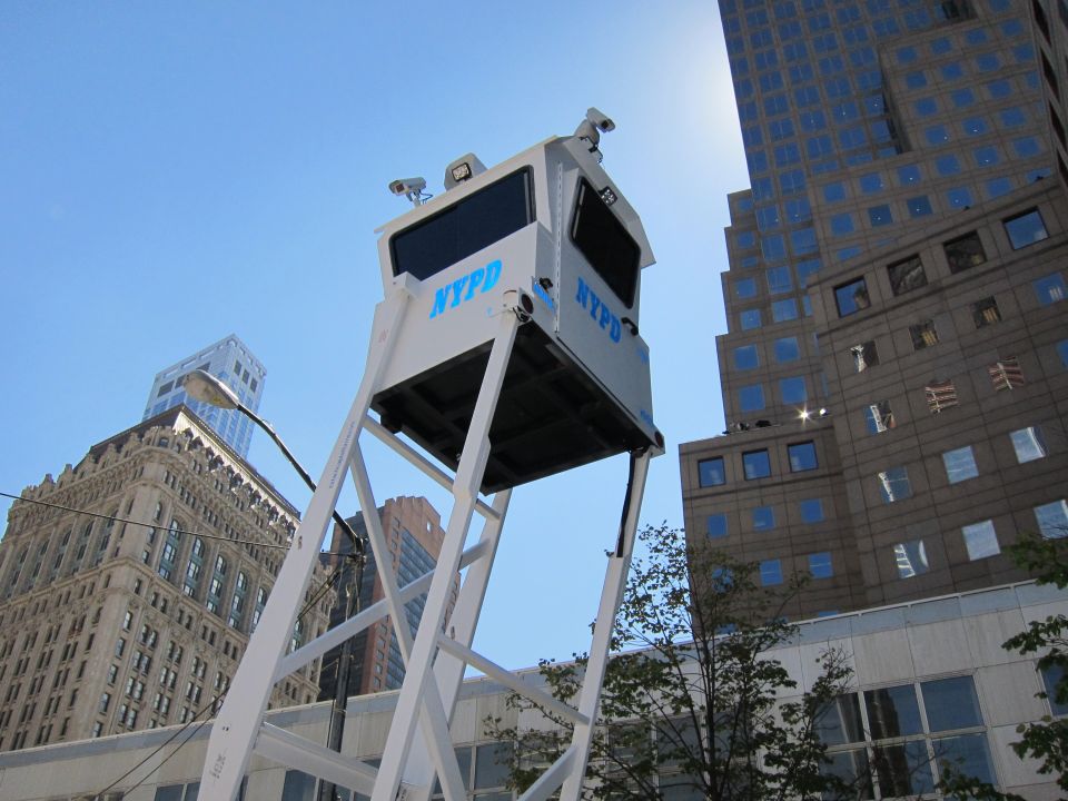 White NYPD observation tower on tall legs against blue sky and skyscrapers