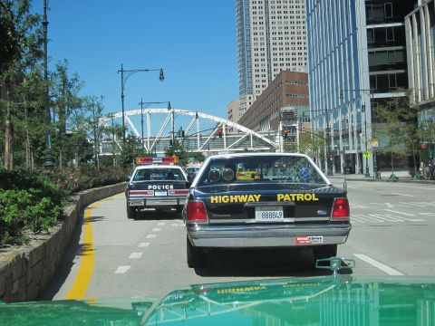 1991 Ford LTD Crown Victoria - black Ford Highway Patrol sedan with plate 888849 drives behind a white police car on a downtown street near a bridge.