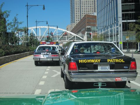 1991 Ford LTD Crown Victoria - rear of a black Ford Highway Patrol cruiser, plate 888849, is shown trailing a white police sedan on a sunny city street.