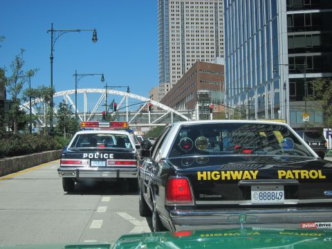 1991 Ford LTD Crown Victoria - black Ford Highway Patrol sedan and a white police car travel a downtown street with a pedestrian bridge behind.