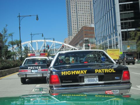 1991 Ford LTD Crown Victoria - black Ford Highway Patrol cruiser, plate 888849, follows a white police car down a city avenue near tall buildings.