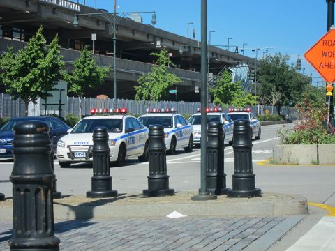 1991 Ford LTD Crown Victoria - line of white NYPD patrol cars sits along a street near black bollards with an elevated highway and Road Work Ahead sign behind.