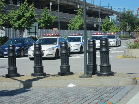 1991 Ford LTD Crown Victoria - White NYPD patrol cars and a blue sedan are parked across a street behind a row of black ornamental bollards near an overpass.