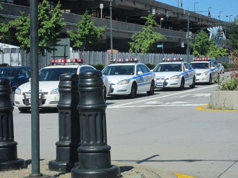 1991 Ford LTD Crown Victoria - Several white NYPD patrol cars are lined up along a roadway beyond black bollards, with an elevated highway in the background.