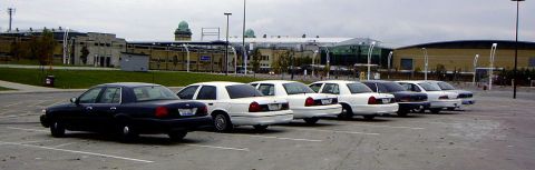 1999 Ford Crown Victoria - A dark blue 1999 Ford Crown Victoria is parked in a lot alongside several white sedans, with a building in the background.