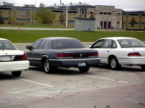 1999 Ford Crown Victoria - dark green Mercury Grand Marquis sedan parked between two white Ford sedans in a lot near a building.