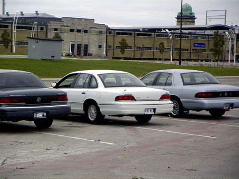 1999 Ford Crown Victoria - Three sedans are parked in a lot with a large building in the background.