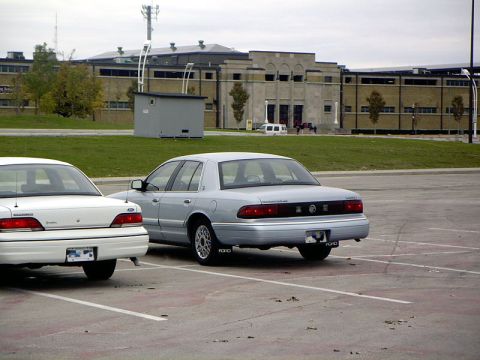 1999 Ford Crown Victoria - Two sedans are parked in a lot with a building in the background.