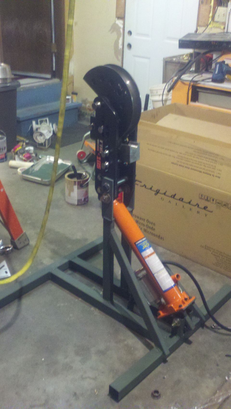 Hydraulic metal bender with orange jack on a green stand, in a workshop
