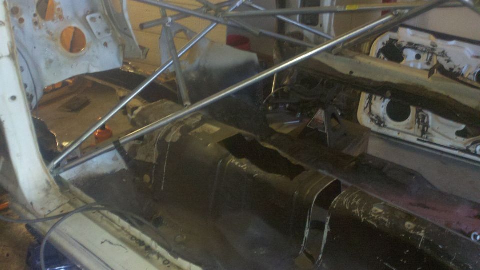 Car interior stripped down with roll cage installed, showing floor pan and structural supports