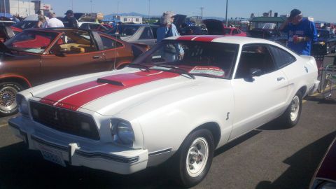 1978 Ford Mustang II - A white 1978 Ford Mustang II with red racing stripes is parked at an outdoor event with other vehicles.