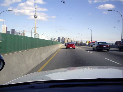 1999 Ford Crown Victoria - A red sedan is seen driving on a highway with the Toronto skyline visible in the distance under a partly cloudy sky.