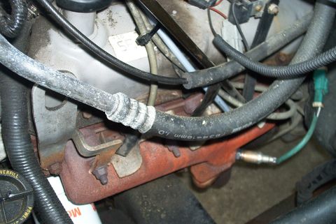 1996 Ford Bronco - Close-up of an engine bay showing a rusty exhaust manifold with a downstream oxygen sensor installed, plus a corroded rubber hose stamped '…