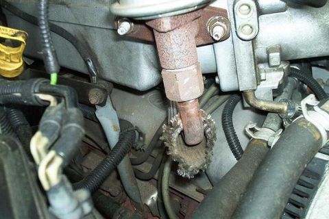 1996 Ford Bronco - Close-up down into an engine bay at a heavily rust-scaled steel tube with a hex fitting and a frayed, disintegrating braided heat shield or…