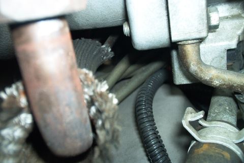 1996 Ford Bronco - Tight close-up deep in an engine bay showing a rusty exhaust manifold tube wrapped in frayed heat-shield braid, with black corrugated wirin…