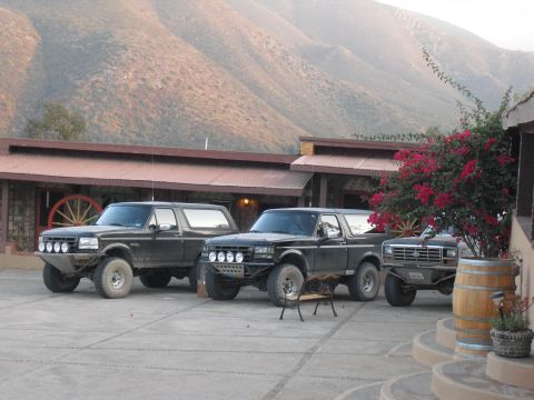 1986 Ford Bronco - Three modified Ford Broncos are parked in a courtyard with a building and mountains in the background.