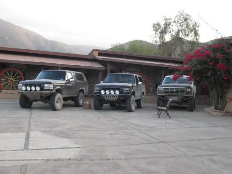 1986 Ford Bronco - Three modified Ford Broncos are parked in front of a building with a rustic facade and mountains in the background.