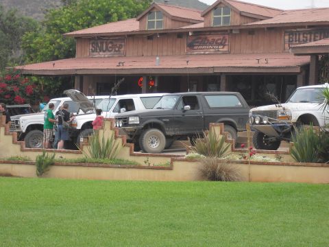 1986 Ford Bronco - Several off-road vehicles are parked in front of a building with various sponsor signs, including Eibach Springs.