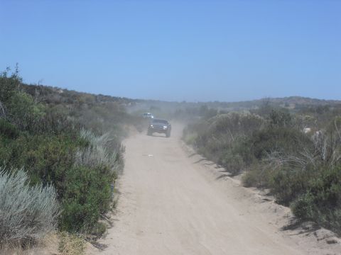 1986 Ford Bronco - Two trucks kick up dust as they drive down a dirt road through a dry, scrubby landscape under a clear blue sky.