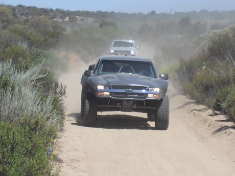 1986 Ford Bronco - Two trucks traverse a dusty desert road, one in the foreground and another following behind.