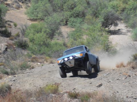 1986 Ford Bronco - A blue off-road truck navigates a dusty, terrain, kicking up a cloud of dust as it drives.