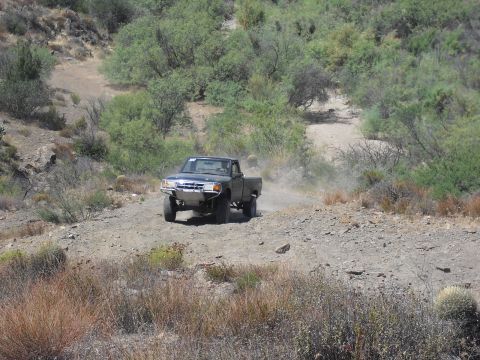 1986 Ford Bronco - A pickup truck kicks up dust as it navigates a rough, unpaved road through a dry, scrub-covered landscape.