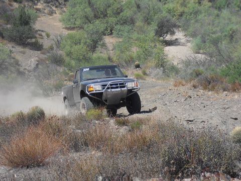 1986 Ford Bronco - A black Ford Ranger pickup truck navigates a dusty, off-road trail with desert vegetation.