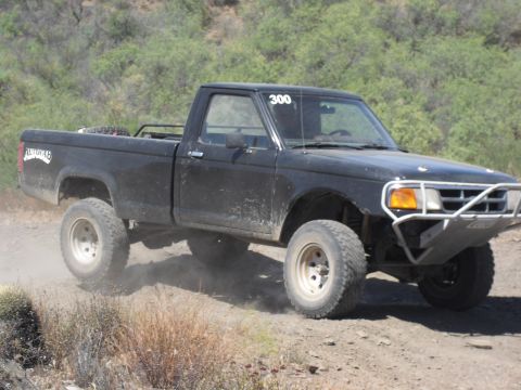 1986 Ford Bronco - A black pickup truck modified for off-road racing navigates a dusty trail.